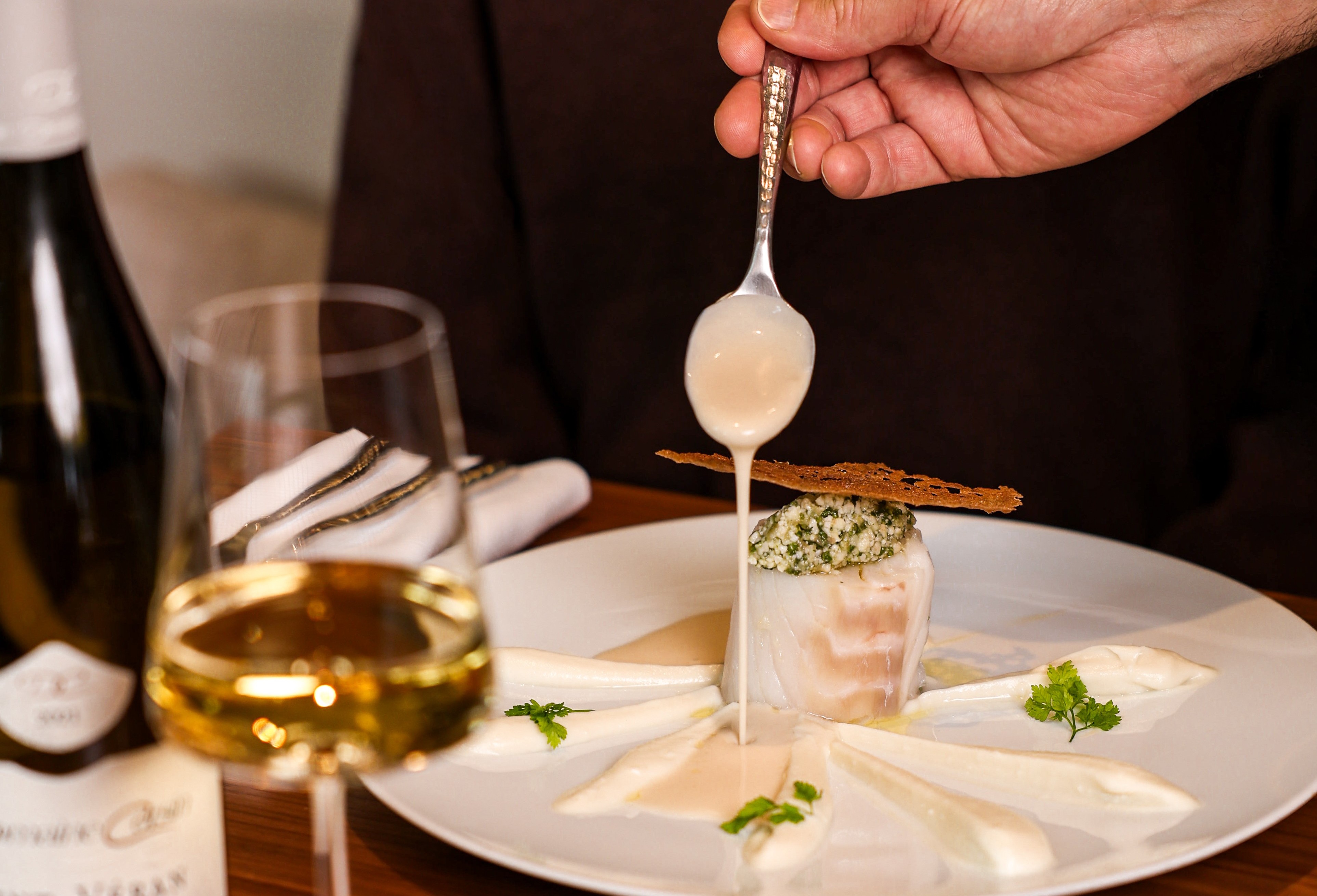 Dressage d’une assiette gastronomique au restaurant Epona de l’InterContinental Lyon – Hotel Dieu, avec une sauce délicatement versée sur un plat raffiné, accompagné d’un verre de vin blanc.